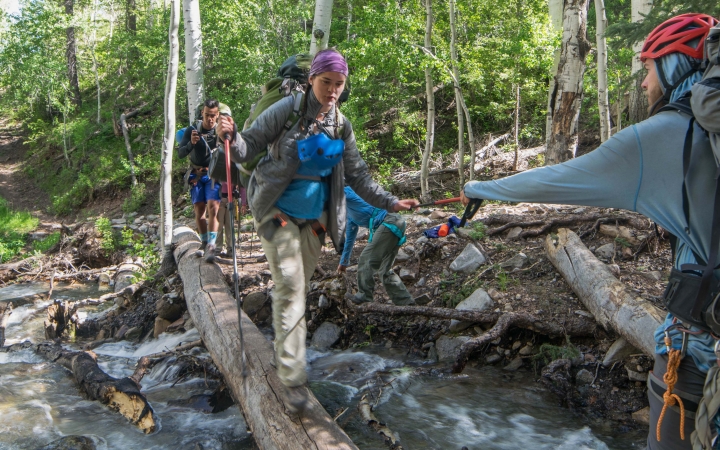 A person crosses a creek over a log while another reaches out their arm to support them. 